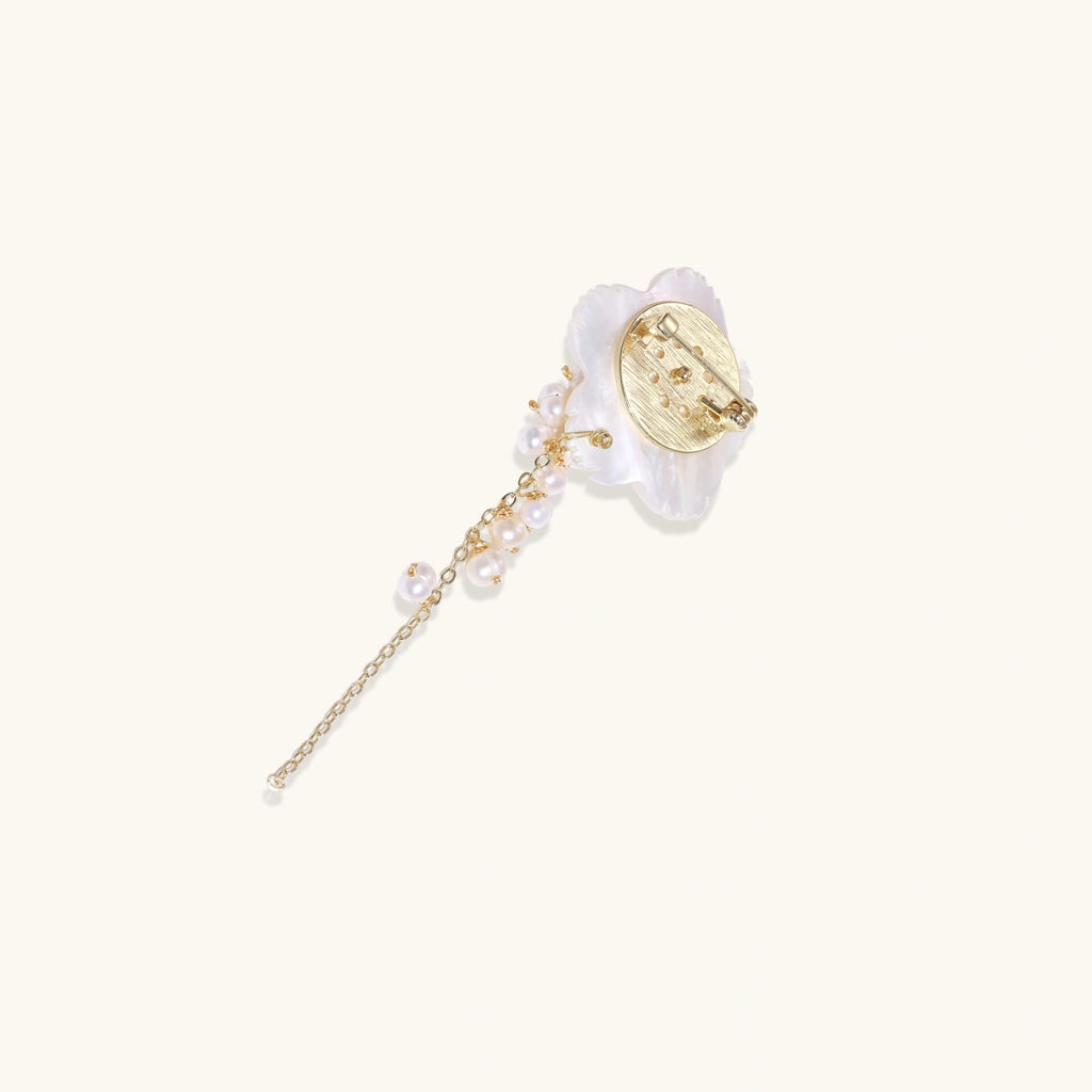Iridescent shell flower brooch with carved petals and cascading freshwater pearls on gold chain against cream background.
