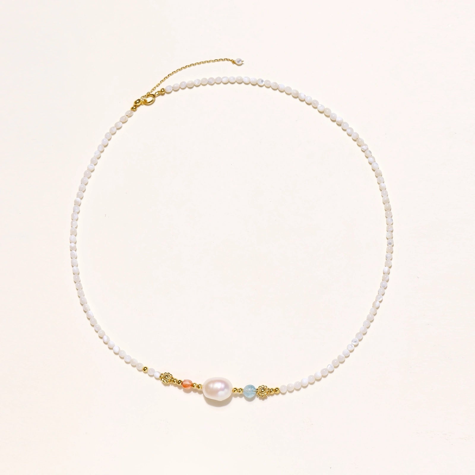 Delicate white beaded necklace with baroque pearl center, gold accents, and coral and aqua gemstones on cream background.